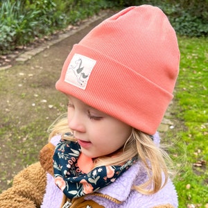 May include: A young child wearing a coral pink beanie with a small white unicorn graphic. The beanie has a ribbed texture. The child is also wearing a black and floral patterned scarf.