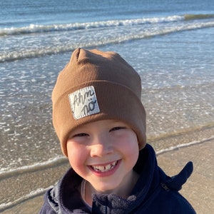 Könnte beinhalten: Ein kleines Kind trägt eine braune Mütze mit einem weißen Aufnäher, auf dem "ahm no" steht, und lächelt in die Kamera. Das Kind trägt eine blaue Fleecejacke und steht an einem Sandstrand.