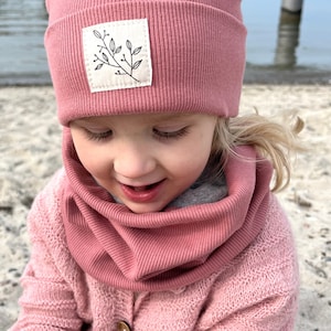 Könnte beinhalten: Eine rosafarbene gerippte Strickmütze mit einem kleinen weißen Patch, auf dem ein schwarzer Zweig mit Blättern abgebildet ist. Die Mütze wird von einem Kind mit blonden Haaren getragen.