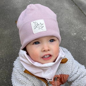 Op de afbeelding: Een roze gebreide beanie met een witte patch met een eenvoudige lijntekening van een tak met bladeren. De beanie wordt gedragen door een kind met blauwe ogen.