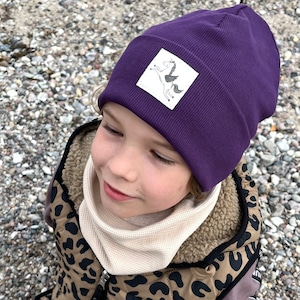 May include: A deep purple beanie with a white square patch featuring a unicorn illustration. The beanie is ribbed and appears to be made of a soft material. The child is wearing a beige neck warmer and a leopard print jacket.