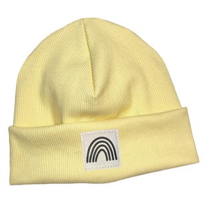 May include: A pale yellow beanie with a ribbed texture. A white square patch with a black rainbow design is sewn on the front. The beanie has a folded brim. The word "kinnerleev" is written in cursive at the bottom.
