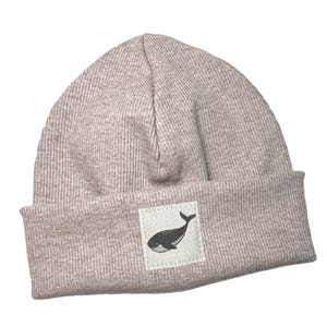 Könnte beinhalten: Hellmauve gerippte Strickmütze mit umgeschlagenem Saum. Auf der Vorderseite ist ein kleines quadratisches Patch mit einer schwarzen Walillustration aufgenäht. Das Wort "kinnerleev" steht unten in Schreibschrift.