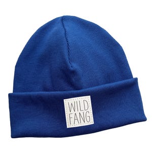 Può includere: Un berretto blu reale con bordo ripiegato. Una toppa quadrata bianca con la scritta "WILD FANG" è cucita sul davanti. Il cappello è realizzato in tessuto a maglia a coste.