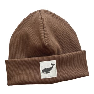 Könnte beinhalten: Braune Beanie-Mütze mit umgeschlagenem Saum. Ein kleines quadratisches Patch am Saum zeigt eine schwarz-weiße Wal-Illustration. Die Mütze ist aus geripptem Strickstoff gefertigt.