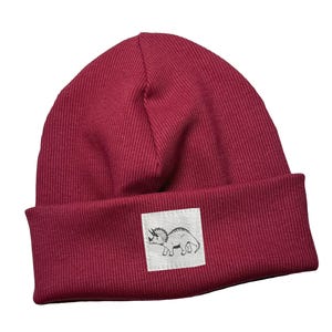 May include: A deep red ribbed knit beanie hat with a turned-up brim. A small white square patch on the front displays a black and white triceratops illustration. A fashionable winter accessory.