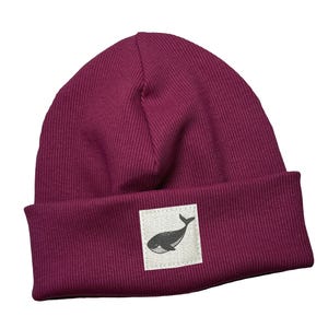 May include: A burgundy ribbed knit beanie with a folded brim. A square patch with a black whale illustration is sewn on the front. The word "kinnerleev" is written in cursive script in the lower right corner.