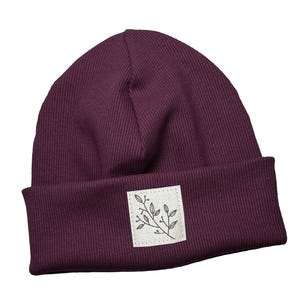 Può includere: Un berretto in maglia a coste bordeaux scuro con risvolto. Una piccola toppa quadrata con un'illustrazione botanica è cucita sul davanti. Un accessorio alla moda per l'inverno.