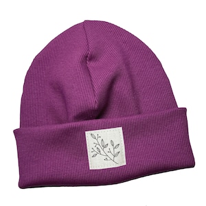 Può includere: Un berretto in maglia a coste viola scuro con bordo ripiegato. Una piccola toppa quadrata con un'illustrazione botanica è cucita sull'orlo. Un accessorio elegante per il freddo.