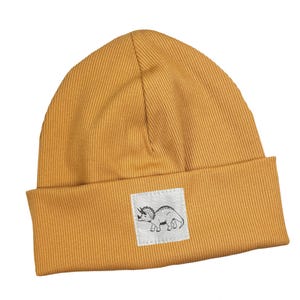 May include: Mustard yellow ribbed knit beanie with a folded brim. A small square patch features a black and white illustration of a triceratops. The word "kinnerleev" is written in cursive script.