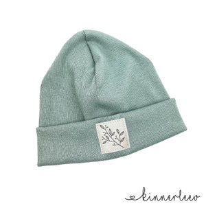 Op de afbeelding: Een lichtblauwe beanie met een klein wit patch met een zwarte lijntekening van een tak met bladeren. De beanie is gemaakt van een zachte, geribbelde stof.