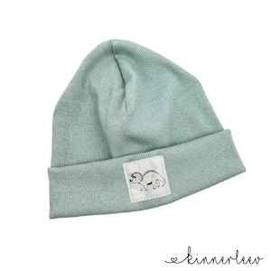 Könnte beinhalten: Eine hellblaue Strickmütze mit einem weißen Patch, auf dem ein schwarz-weißes Bild eines Dinosauriers zu sehen ist.