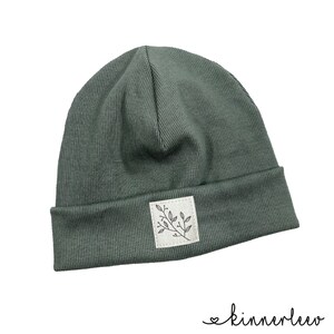 May include: A dark green beanie with a small white patch featuring a simple black line drawing of a branch with leaves. The beanie is made of a ribbed fabric.