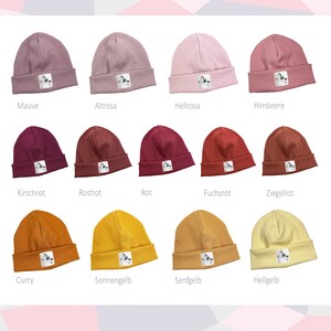 Könnte beinhalten: Ein Set mit 10 bunten Strickmützen in verschiedenen Farbtönen von Rosa, Rot, Gelb und Orange. Jede Mütze hat einen kleinen weißen Aufnäher mit einem schwarzen Design.