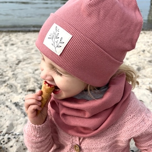 May include: A young child wearing a pink ribbed beanie with a white leaf design patch and a matching pink ribbed neck warmer. The child is eating a waffle cone.