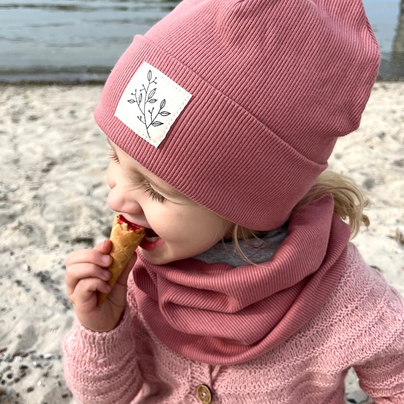 May include: A young child wearing a pink ribbed beanie with a white leaf design patch and a matching pink ribbed neck warmer. The child is eating a waffle cone.