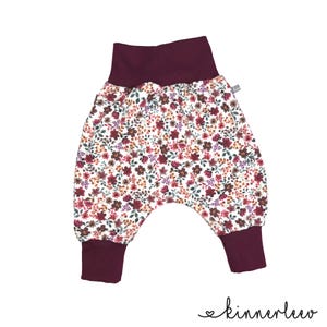Op de afbeelding: Broek met verlaagd kruis op een witte achtergrond met een dicht bloemenpatroon in bordeaux, roze en groen. Het kledingstuk heeft een brede, effen bordeauxrode tailleband en bijpassende bordeauxrode boorden bij de enkels. Tekst leest kinnerleev.