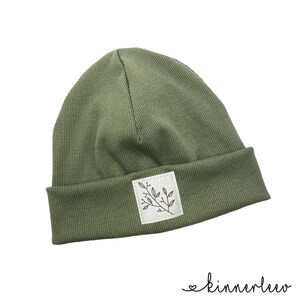 Puede incluir: Un gorro de punto verde con un peque&ntilde;o parche blanco que presenta un dibujo de l&iacute;nea negra de una rama con hojas. El parche tiene un borde cosido.