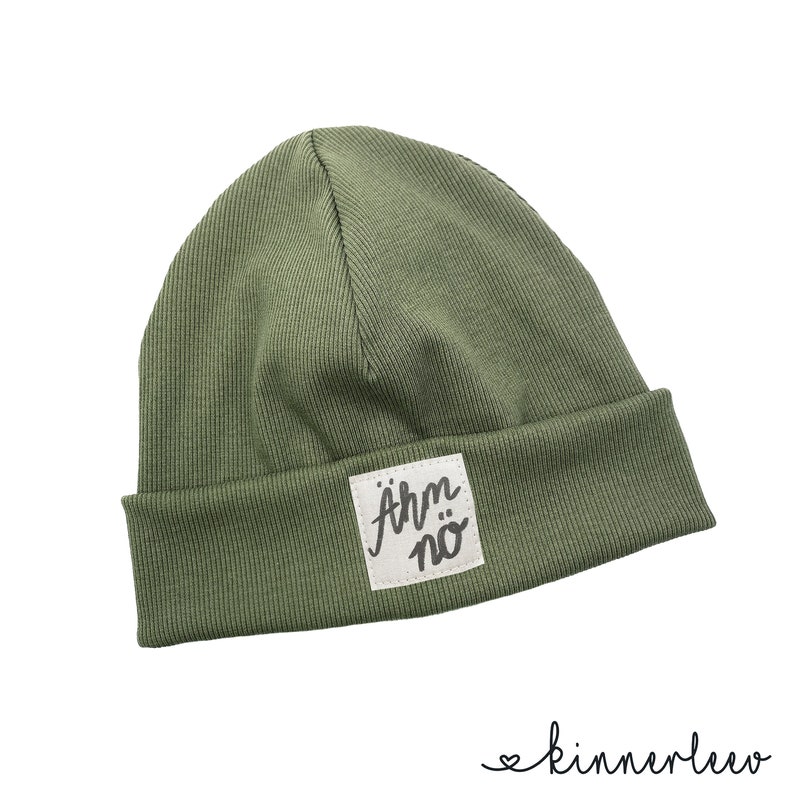 Puede incluir: Un gorro de punto verde con un parche cuadrado blanco que dice "&auml;rm n&ouml;" en letras negras.