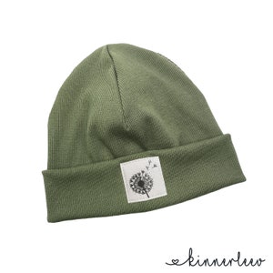 Puede incluir: Un gorro de punto verde con un parche blanco con un dise&ntilde;o de diente de le&oacute;n negro.