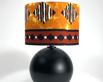May include: A black table lamp with a round base and a drum shade featuring a geometric pattern in shades of orange, brown, white, and black.