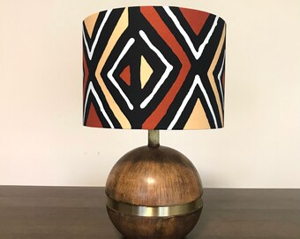 May include: A decorative table lamp with a round wooden base and a patterned lampshade. The lampshade features a geometric design in black, white, tan, and rust colors. The base has a brass-colored band around the middle.