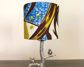 May include: A decorative table lamp with a silver tree trunk base and two small bird figurines perched on branches. The lampshade features a colorful patterned fabric with blue, yellow, and brown designs. The lamp is designed for indoor use.