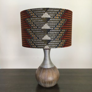 Op de afbeelding: Een tafellamp met een bruine houten voet en een zilveren metalen steel. De lampenkap is gemaakt van stof met een geometrisch patroon in tinten zwart, oranje, rood, wit en blauw.