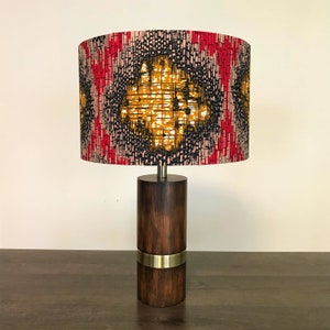 May include: A table lamp with a wooden base and a cylindrical lampshade. The lampshade is made of fabric with a geometric pattern in shades of red, black, and yellow.