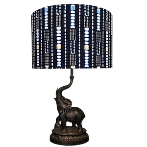 May include: A table lamp with an elephant-shaped base in bronze tones, supporting a cylindrical lampshade. The lampshade has a dark blue background with a repeating pattern of white and gold vertical lines and dots. The lamp is designed for indoor use.