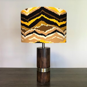 Op de afbeelding: Een tafellamp met een bruine houten voet en een zilveren band. De lampenkap is geel, bruin en zwart met een chevron-patroon.