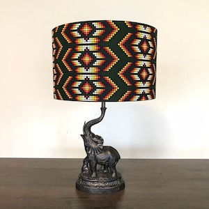 May include: A table lamp with a dark green lampshade displaying a geometric pattern in red, yellow, and white. The lamp base is a bronze-coloured elephant sculpture. The lamp is on a dark wooden surface.