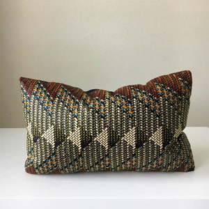 May include: Rectangular decorative pillow with a woven pattern in black, white, orange, blue, and red. The pillow has a textured appearance and is placed on a white surface.