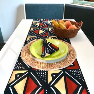 Peut inclure: Un chemin de table &agrave; motifs g&eacute;om&eacute;triques noirs, rouges et jaunes avec une assiette verte et un dessous de plat en osier. Le chemin de table est d&eacute;cor&eacute; de points blancs et une serviette &agrave; motifs noirs et blancs est pli&eacute;e sur l'assiette.