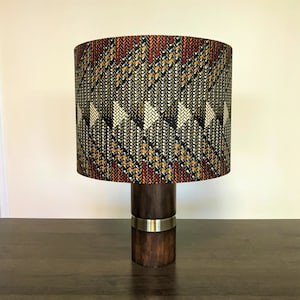 Può includere: Una lampada da tavolo con una base in legno scuro e un paralume con motivo. Il paralume presenta un motivo geometrico in tonalità di nero, bianco, arancione, rosso e blu.