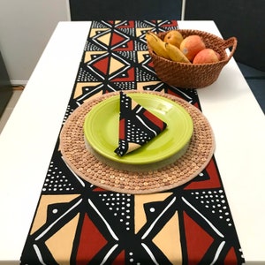 Peut inclure: Un chemin de table noir, rouge et beige avec un motif g&eacute;om&eacute;trique. Le chemin de table est sur une table blanche avec une assiette verte et un dessous de plat en osier. Un panier en osier avec des fruits est sur la table.