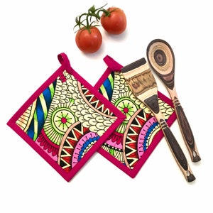 May include: Two colorful square pot holders with a pink border and a geometric pattern. The pot holders are made of fabric and are quilted. They are lying on a white surface with two wooden spoons.