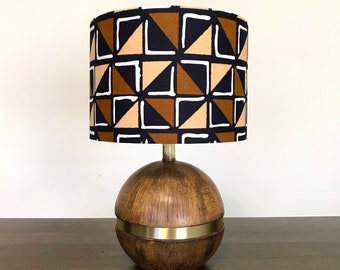 May include: A wooden table lamp with a round, brown base and a gold band. The lampshade is white with a geometric pattern of black, brown, and beige triangles.