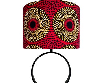 Peut inclure: Une lampe de table avec un abat-jour cylindrique présentant un motif géométrique circulaire rouge, noir et jaune. La base de la lampe se compose d'un fin anneau en métal noir soutenu par un pied carré noir, créant un luminaire moderne.