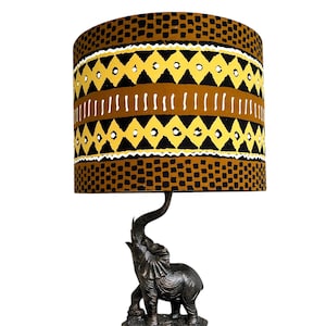 May include: A table lamp featuring a bronze-colored elephant base with its trunk raised upward. The cylindrical lampshade displays a repeating pattern of black squares, yellow diamonds, and white vertical lines on a brown background.