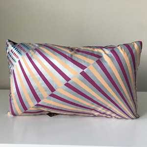 Könnte beinhalten: Ein rechteckiges Kissen mit einem geometrischen Muster. Das Muster besteht aus abwechselnden Streifen in Pink, Beige und Silber. Das Kissen hat einen schwarzen Rand.
