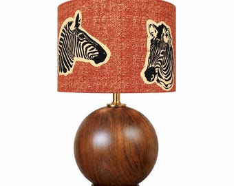 Könnte beinhalten: Eine Tischlampe mit einem kugelförmigen Sockel aus dunkelbraunem Holz und einem zylindrischen Lampenschirm. Der Schirm besteht aus strukturiertem orangefarbenem Stoff mit zwei schwarz-weißen Zebra-Illustrationen, eine im Profil und eine von vorne.