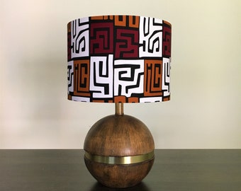 May include: A wooden table lamp with a round, brown base and a gold band. The lampshade is white with a geometric pattern in orange, brown, and red.