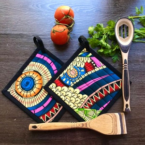 May include: Two patterned pot holders with black trim and hanging loops. A wooden slotted spoon and spatula are next to the pot holders. Two tomatoes on a vine and parsley are also in the image. The pot holders have a geometric design with blue, pink, and yellow.