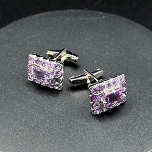 May include: A pair of silver cufflinks with rectangular faces. Each cufflink is adorned with a central, rectangular purple stone, surrounded by smaller, round purple stones. The cufflinks are set against a dark background.