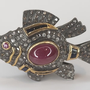 May include: A silver and gold fish-shaped ring with a large red gemstone and smaller diamonds. The ring is detailed with black gemstones and has a textured finish.