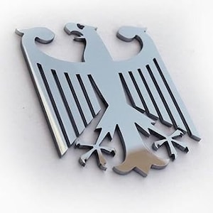 May include: A silver metal eagle emblem with a stylized design. The eagle has its wings spread and is holding a cross in its talons.