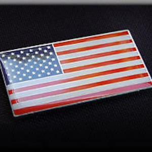 May include: A small, rectangular American flag patch with a red, white, and blue design. The flag is made of a shiny, reflective material.