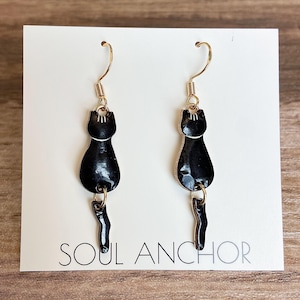 Puede incluir: Un par de pendientes de gato negros con ganchos y detalles dorados. Los pendientes presentan una silueta de gato negro brillante con cola. El texto "SOUL ANCHOR" está impreso en la tarjeta blanca detrás de los pendientes.