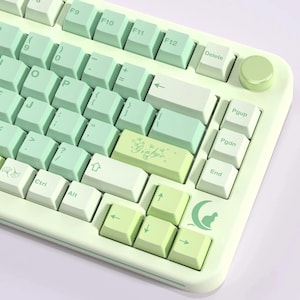 May include: A pastel green and white mechanical keyboard with gradient keycaps. The keyboard features a variety of keys, including function keys, arrow keys, and a delete key. The keycaps have a light green and white color scheme, with some keys featuring small illustrations.
